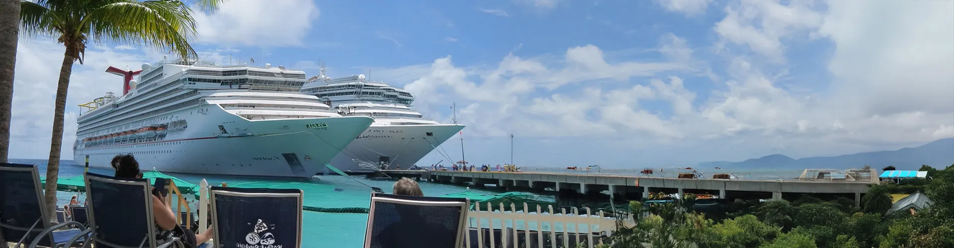 Cruise ships docked at a tropical port with palm trees and beach chairs in the foreground