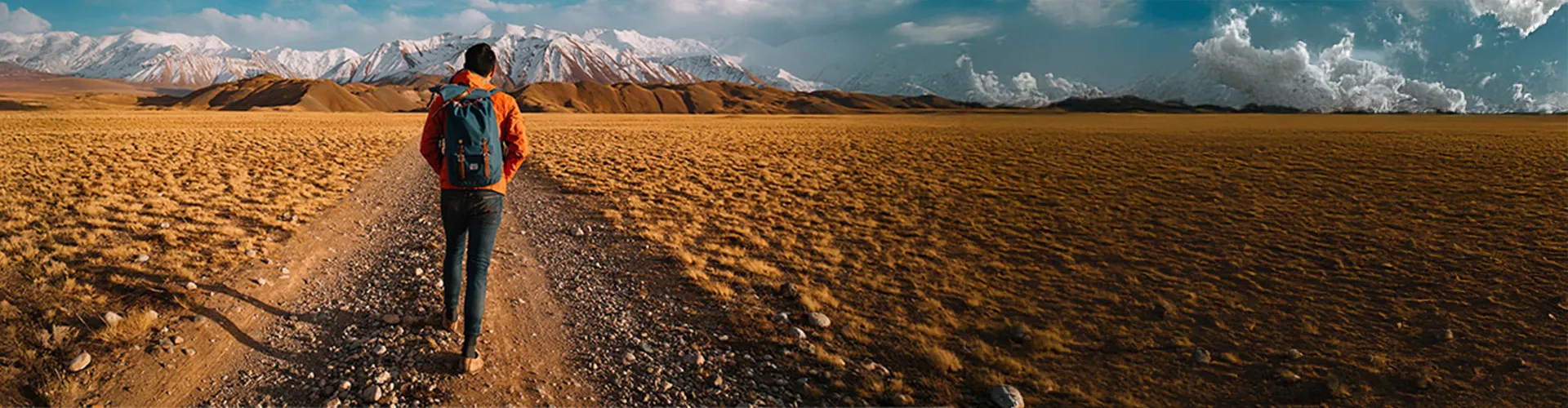 Traveler with backpack walking along a gravel path through a vast open landscape with snow-capped mountains